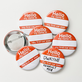 Six round pinback buttons with a white and orange design reading 'Hello my name is my first car was' are arranged on a light background, with one button showing the metal pin-back side.